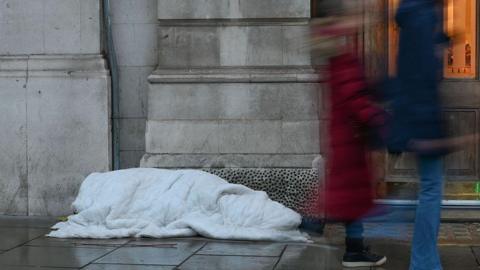 A homeless person underneath a white blanket on a wet street. Two blurred people are walking past