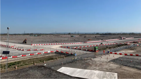 A large stretch of land is under construction. There are red and white bollards and gravel on the ground.
