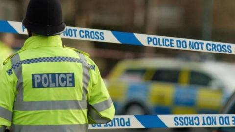 Police officer wearing helmet  in hi vis with back to camera with police tape and police car behind him 