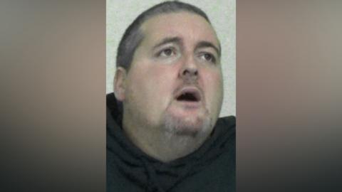 Mugshot of Michael Gill. He has a large head and short dark grey hair. He is looking up and to his left with his mouth wide open and a confused look in his eyes.