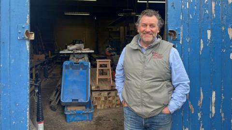 A man is standing at the blue wooden gated entrance to a workshop, with his hands in his pockets. He is wearing jeans a grey/green gillet and a pale blue short. He is smiling at the camera. He has grey curly hair and is unshaven. 