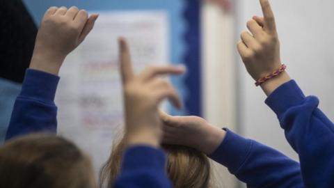 Children in a classroom are pictured from behind. They all have their hands raised in the air as if to answer a question and are wearing blue jumpers. 
