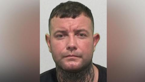 Mugshot of Hazard. He has a large tiger's face tattooed across the entirety of the front of his neck, is clean shaven with short dark hair.