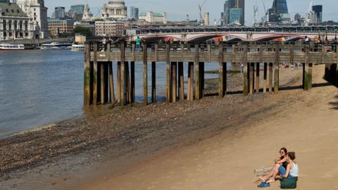 Two young women pictures sitting on a beach by the river Thames, with the London skyline in the background