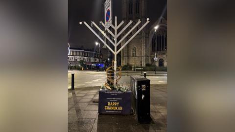 A large decorative silver Hanukkah menorah on a street, on a stand which reads 'Happy Chanukah'. The lightbulbs representing candles on the top of the menorah have been damaged.