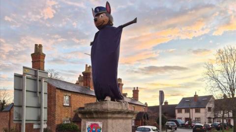 A statue dressed as a dog character Chase from children's TV programme Paw Patrol. The statue is in a village square, with houses visible in the background and cars parked on the street. The German Shepherd dog is wearing a blue  sheet and his arms are out wide.