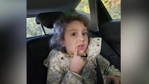 Maisie is sat in the back seat of a car. She has curly brown hair and is wearing a pale flowery rain coat. She has her right hand index finger in her mouth, pointing at her small tooth. 