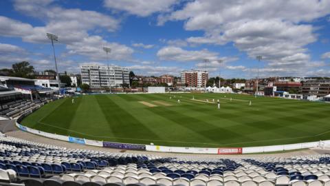 A cricket pitch with players on it. A number of seats are set up around the perimeter.