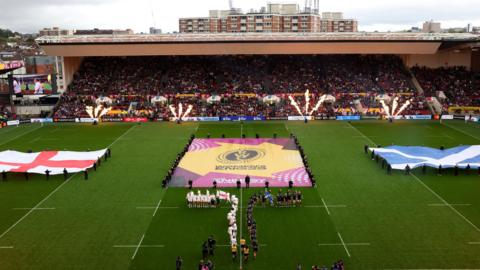 The ground at Ashton Gate Stadium on a cloudy and wet day. There are people holding a large England flag and a large Scotland flag on the pitch on either side, with Women's Rugby World Cup branding in the middle. The players can be seen filing out onto the pitch as fans pack the stands.