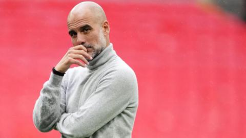 Pep Guardiola wearing a grey turtle neck against a red background. He looks thoughtful.