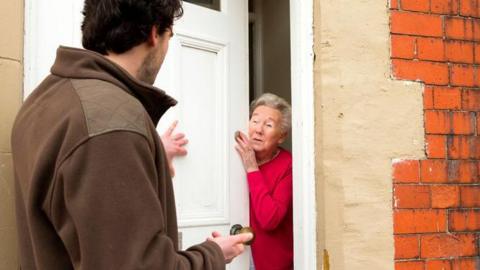 An older woman in a red jumper opens her front door to a man. He is gesturing and putting her under pressure.