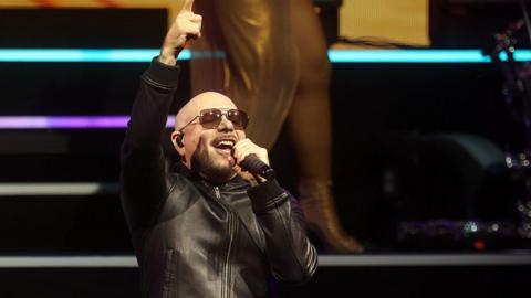 Pitbull, a bald man wearing aviator sunglasses, singing on stage. He is wearing a black leather jacket.