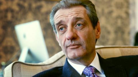 A grey-haired man in a dark suit, wearing a white shirt and a purple tie, sits on a cream sofa in a room, looking slightly away from the camera.