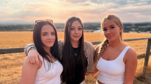 Amelie is on the left, wearing a white top. She has shoulder-length dark fair and sunglasses on top of her head. Liv is in the middle and is wearing a grey bolero top over a black top. She has long black hair. Vik is on the right, wearing a white top. Her blonde hair is in a single plait over her right shoulder.