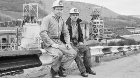 Coal miners Phillip Jenkins (left) and Chris Lee sitting next to each other on road railings at the Taff Merthyr Colliery in south Wales in 1983