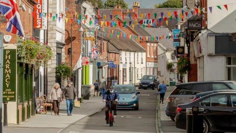 A busy high street in Whitchurch showing people walking, riding bikes. The street is decorated with bunting and England flags. 