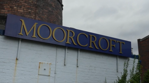 A brick building with the bottom painted white and a blue sign reading "Moorcroft" in gold text.