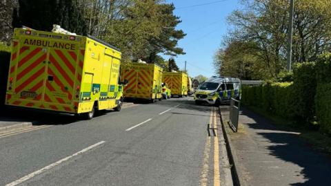 Three ambulances and a police van are parked along a residential road.