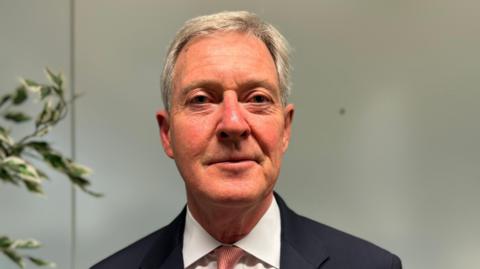 Leader of Surrey County Council, Tim Oliver, looks straight at the camera. He is a white man with grey hair, wearing a white shirt and black blazer. 