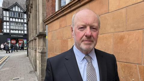 Alan Bates wearing a suit and tie, standing in front of a brown building with shops and shoppers in the background