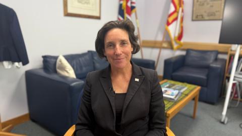 Deputy Lindsay de Sausmarez sits in a office with two flags behind her. She has short brown hair and is wearing a black business suit 