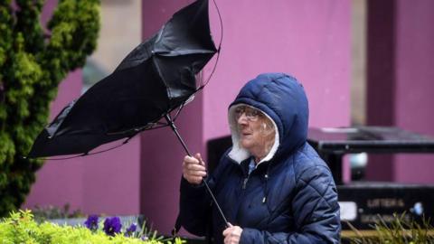 A woman waring a long coat with the hood up is holding an umbrella which has been blown inside-out by high wind as she walks along a street