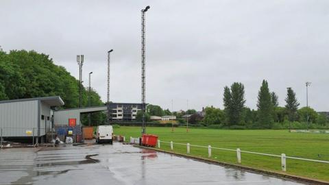 A rugby pitch with car park in the foreground. There is a block of flats in the distance, overlooking the club, and a club house and spectator stand to the left hand side of the image.