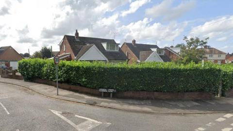 Street view image of the Baylton Drive junction, showing the roofs of large detached houses behind a tall green hedge on the corner of the road.