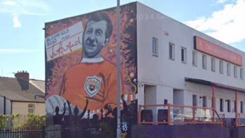 Mural of Jimmy Armfield in Blackpool kit on the side of the Armfield Club which is a flat roofed two-storey building with tangerine and white livery.