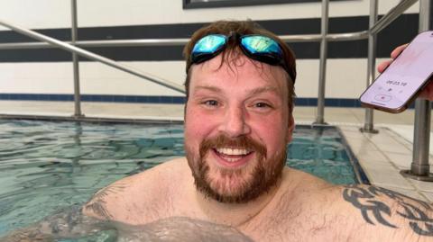 Jordan Brooks smiles in the swimming pool, his goggles on his head. He has brown hair and a beard, with tattoos on his upper arms.