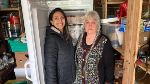 Two women standing side by side in a garage. They are positioned next to an upright freezer with the door open, revealing neatly stacked containers inside. The woman on the left is wearing a dark padded jacket and has long dark hair. The woman on the right is wearing a patterned cardigan with floral detailing and has long grey hair styled in braids. The surrounding area contains various storage items, including plastic bins, boxes, shelves, and household supplies.