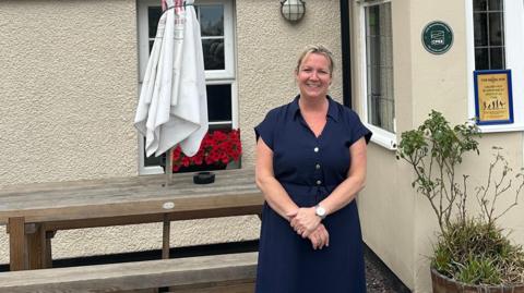 A pub landlady - a middle-aged woman with blonde hair who is wearing a button up navy dress with short sleeves - stands in front of a pub. There is a picnic bench with a parasol behind her as well as some plants.