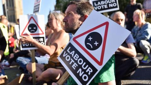 A group of protesters sitting on the ground holding signs that read "Labour vote no," featuring a warning-style symbol with a sad face and crossed-out eyes. The protest is taking place outdoors in daylight, with people in the background and part of the Elizabeth tower visible.