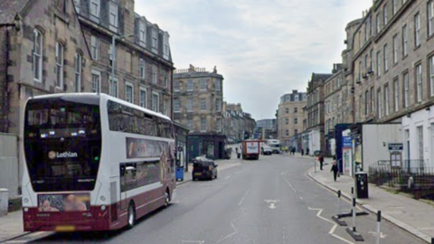 A white and burgundy double decker bus is parked along an old Edinburgh street lined with shops with three storeys of light-coloured flats above them.