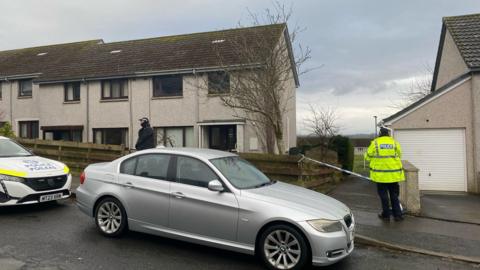 Two police officers and a grey car and a police car sit outside a semi-detached house with the driveway taped off with police tape