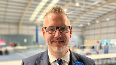 John Campion with short, spikey grey hair. He is pictured in a suit in a sports hall. 