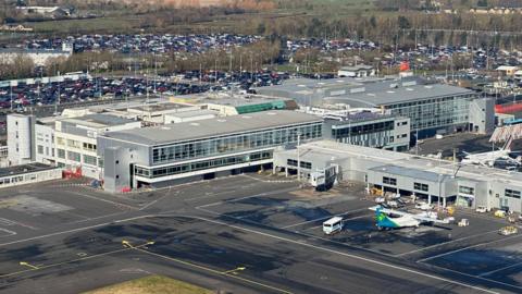 Aerial view of Newcastle Airport. A large plane can be seen on tarmac next to a large terminal building, which is boxy and flat 