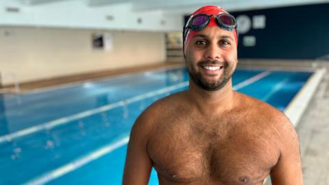 Premal Pattani wearing a red swimming cap and black swimming goggles, smiling at the camera. He is shirtless and standing beside an empty indoor swimming pool, which is out of focus in the background. 
