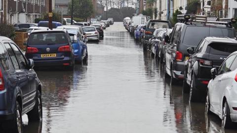 A residential street lined with cars is flooded with dark murky water