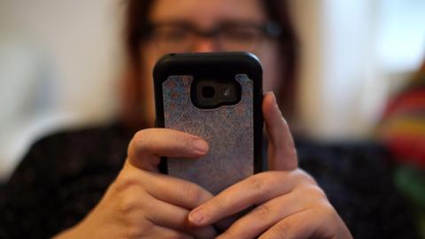 A picture of a lady looking at her phone. She is holding a phone with one camera on the back and a bright phone case.