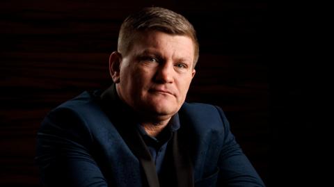 Ricky Hatton, dressed in a navy jacket, and shirt. He has short dark hair and is looking into the camera.