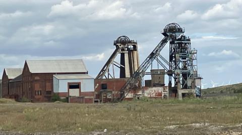 Headstocks at the top of a mine shaft. Next to them is a series of red brick buildings. 