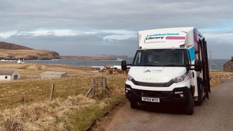 Highlife Highland mobile library van on a narrow road overlooking a rocky bay on a stormy looking day.