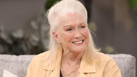 Actress Diane Ladd smiles while sitting on a couch. She is wearing a tan shirt and a gold necklace.