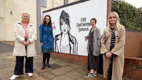 A mural on a white background on a wall showing a young woman with the words Faye Fantarrow forever. Four women stand next to it.