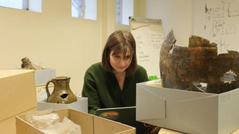 Doctoral Researcher Emily Walker typing on a laptop as she carries out some post-excavation work. She is surrounded by cardboard boxes. Some of the boxes are open, revealing metal and gold looking artifacts inside them. One of the items resembles a vase. Ms Walker is wearing a green cardigan. Her straight brown hair reaches her shoulders and she has a fringe. There is writing on the whiteboards behind her.