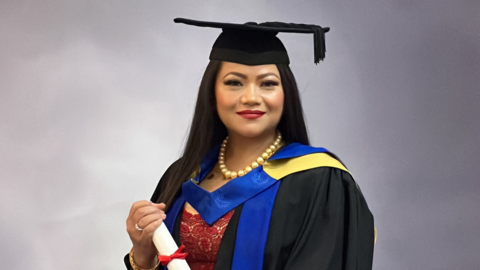 A woman with long black hair dressed in graduation robe and hat holding a diploma