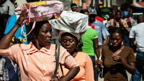 Three Haitian women carry bags on their heads as they walk down a crowded street. Two women wear orange blouses and one wears a brown t-shirt. 