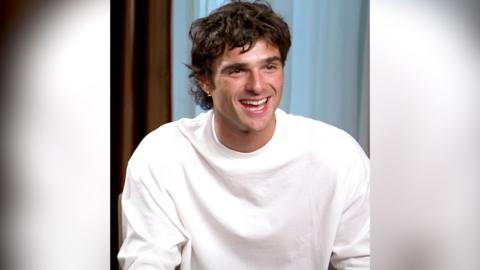 A man, Jacob Elordi, has dark brown hair and wears a white top. He is smiling