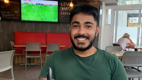 Anugrah Abraham, a man with black hair and black facial hair and a dark green t-shirt smiles directly into the camera. He is sat in a pub, with a TV displaying a football match behind his right shoulder.